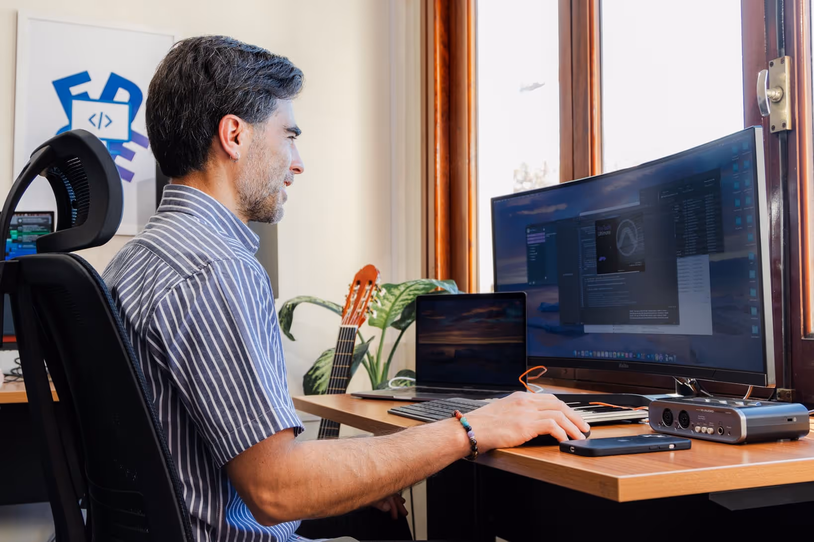 Edge Audio Labs team member focusing on audio software development on a large monitor, using a professional audio interface and a laptop with a guitar nearby, representing the company’s blend of tech expertise and musical passion.